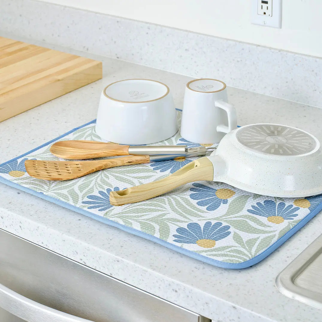 Blue coneflower dish drying mat with a blue and sage leaf pattern, holding white bowls, a pan, and wooden utensils.