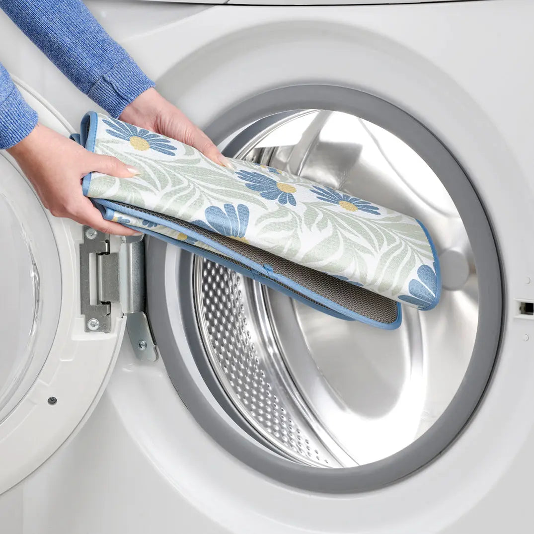 Hands putting a blue and green coneflower print dish drying mat with a gray mesh backing into a washing machine.