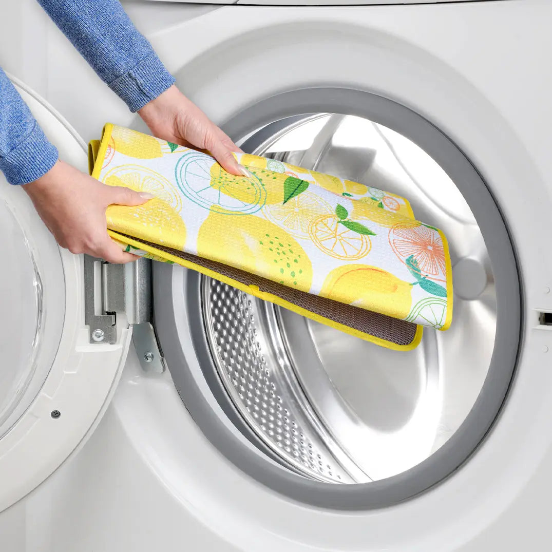 Hands placing a rectangular, yellow lemon-patterned fabric drying mat into a washing machine.