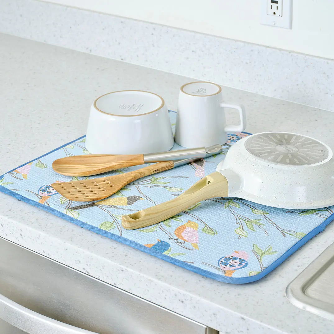 Light blue rectangular dish drying mat featuring a colorful bird and floral pattern, holding kitchenware on a counter.