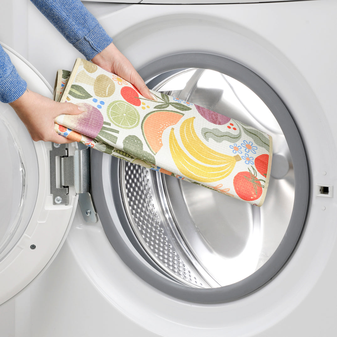 Hands putting a cream-colored, soft-material dish mat with a colorful fruit and vegetable print into a dryer.