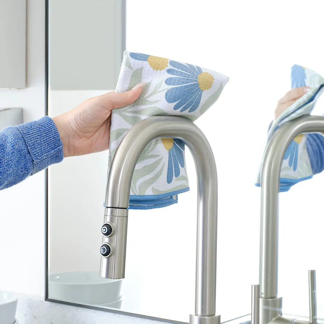 A hand wipes a stainless faucet with a blue and yellow floral coneflower print towel near a mirror.
