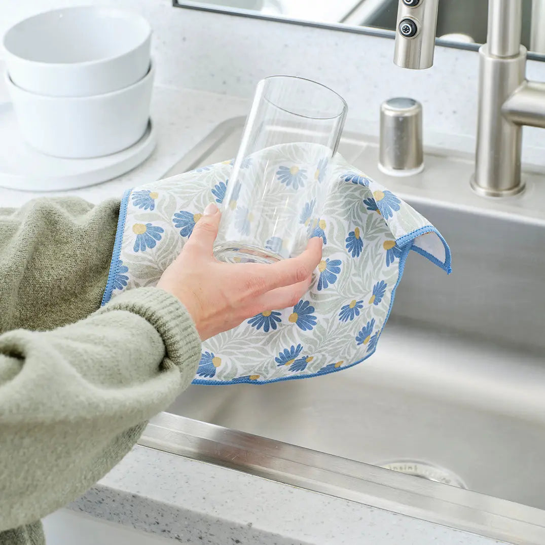 Person drying a clear glass with a light blue and green coneflower-patterned towel over a kitchen sink.