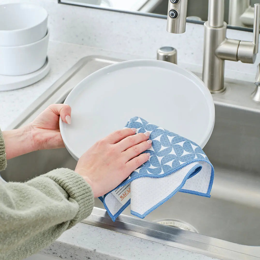 Hands wiping a white plate with a blue and white geometric patterned towel over a stainless steel kitchen sink.