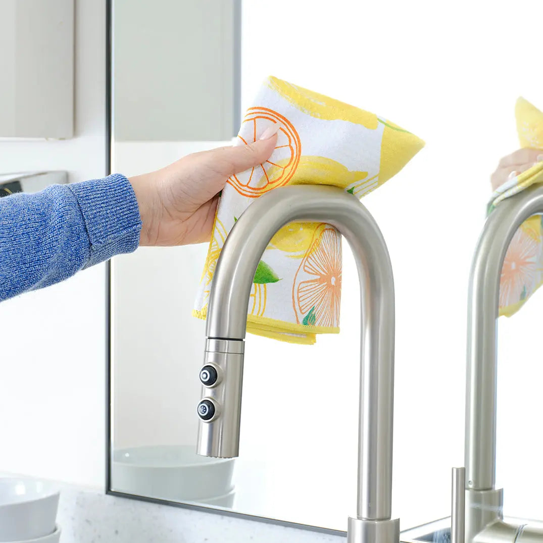 Hand wiping a stainless steel faucet with a white all-purpose towel featuring a yellow and orange lemon print.
