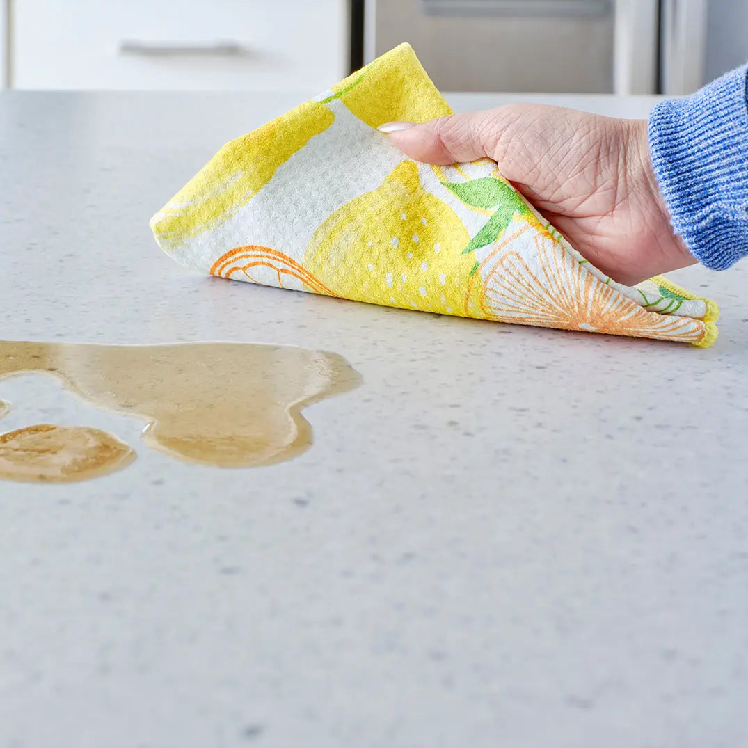Hand wiping a spill on a countertop with a yellow, white, and orange lemon-patterned WerkShoppe all-purpose towel.