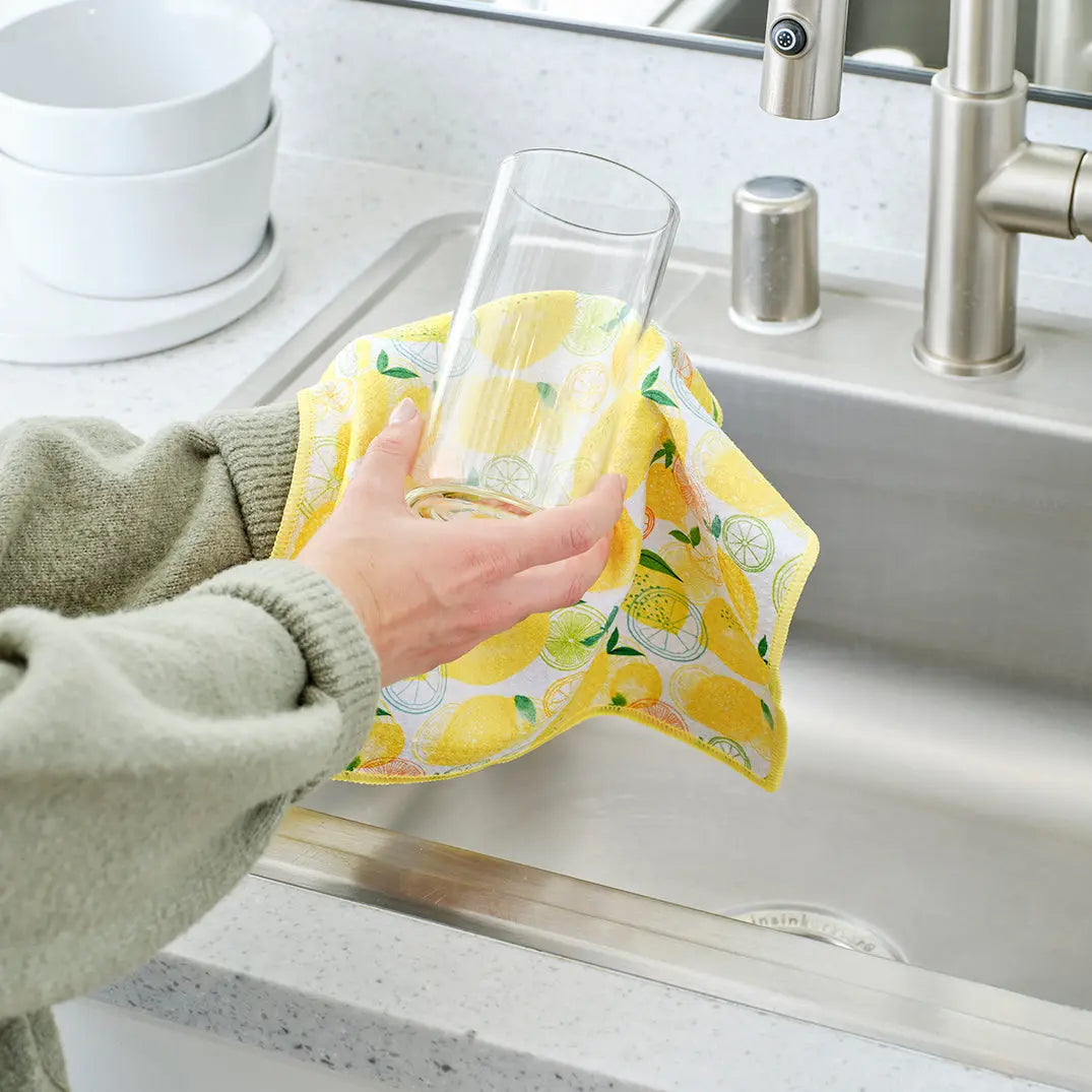 Hand wiping a clear glass with a yellow, white, and green lemon-patterned fabric kitchen towel over a sink.