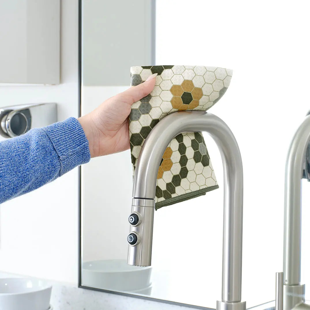 Hand holding an all-purpose towel with a yellow and green honeycomb tile pattern draped over a kitchen faucet.
