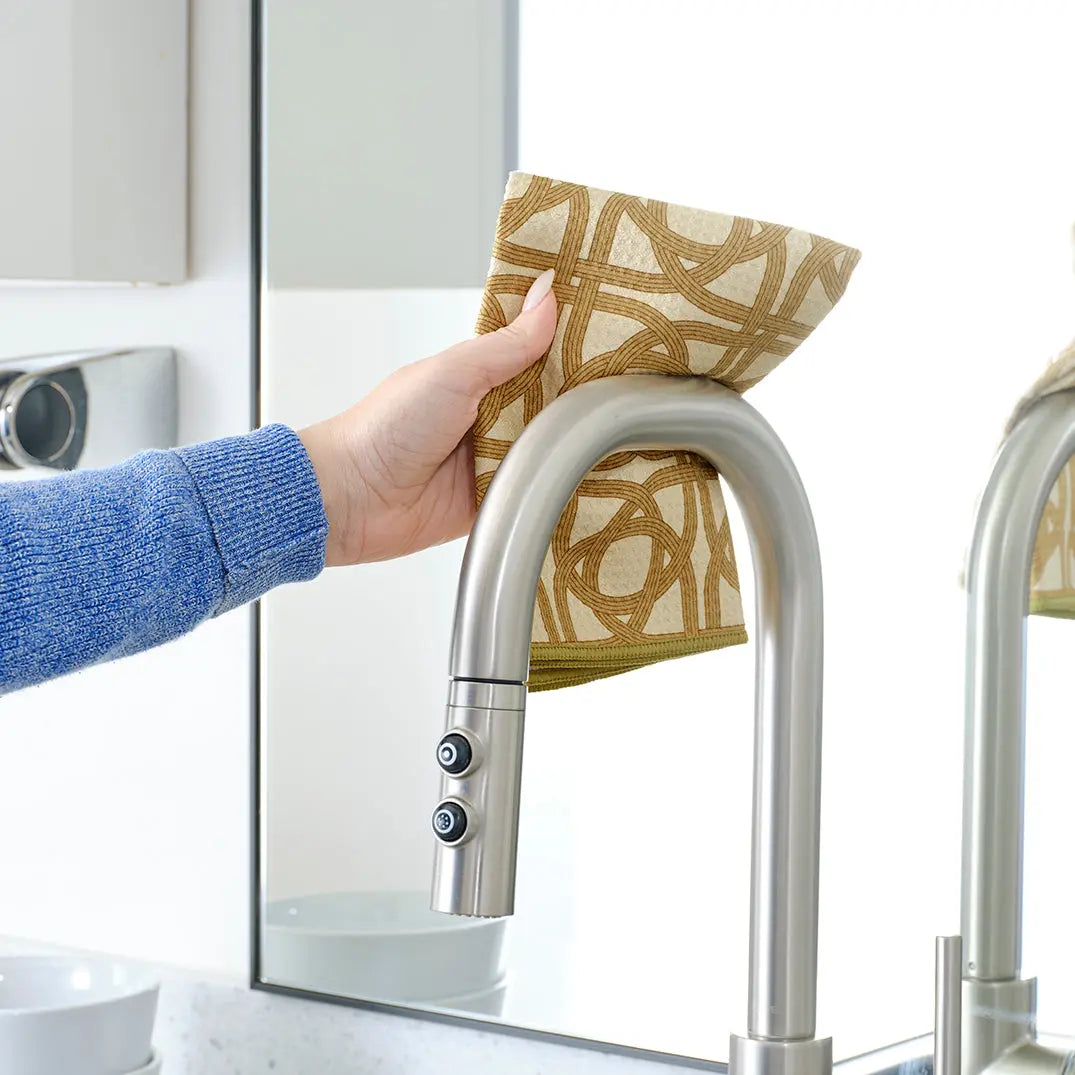 Hand wiping a chrome kitchen faucet with a tan and brown patterned all-purpose towel.