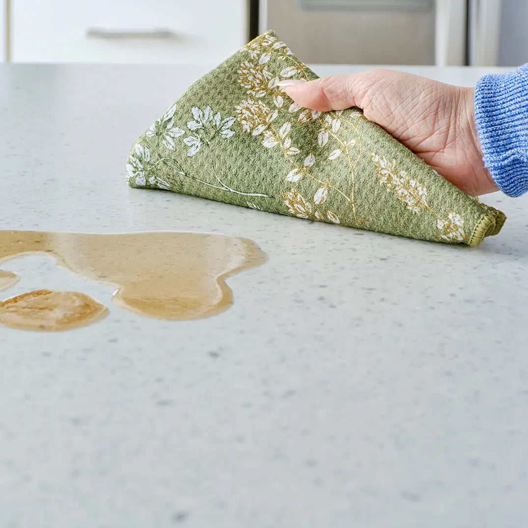 A hand uses a green textured kitchen towel with white herb illustrations to wipe a spill on a speckled countertop.