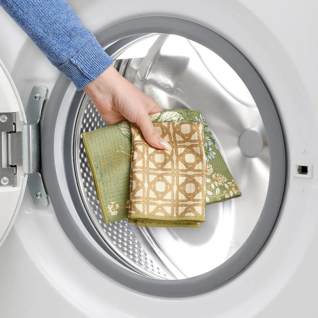 Hand holding kitchen towels with green floral and tan geometric patterns being placed into a laundry machine.