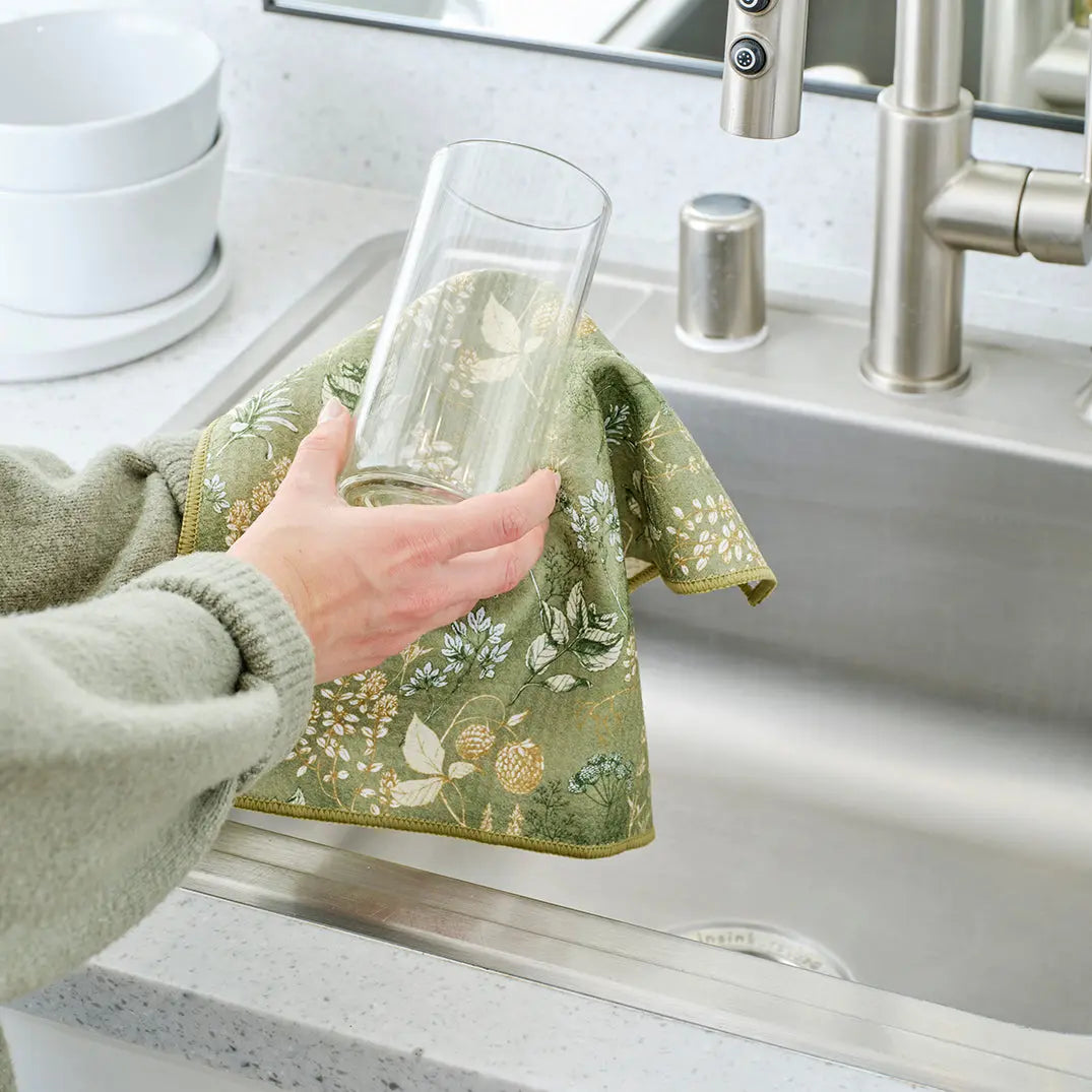 A hand drying a glass with an olive-green kitchen towel featuring a white and yellow botanical herb print.