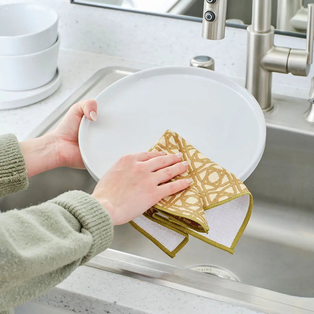 A person wipes a white plate with a tan and white patterned fabric kitchen towel with olive green stitched edges.