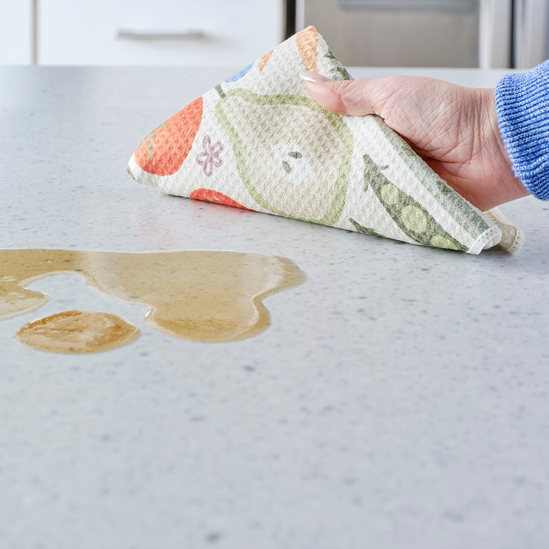 Hand wiping a spill on a countertop with a waffle-texture towel featuring a colorful print of pears and vegetables.