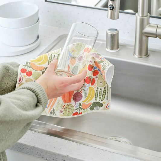Person drying a clear glass with a Farm Stand towel featuring colorful illustrated fruits and vegetables in a kitchen.