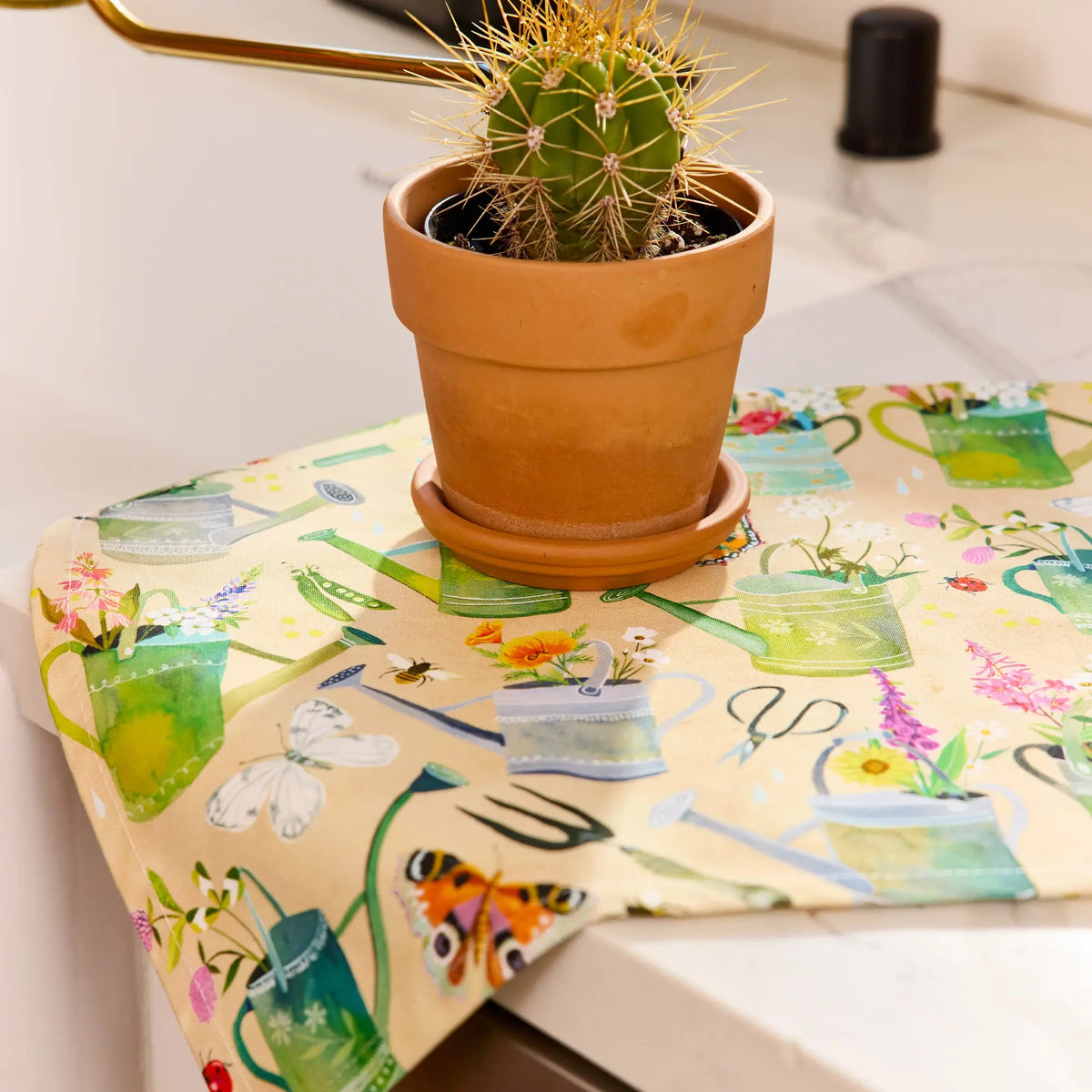 Cotton tea towel with watercolor gardening pattern of watering cans, flowers, butterflies, and tools under a cactus.