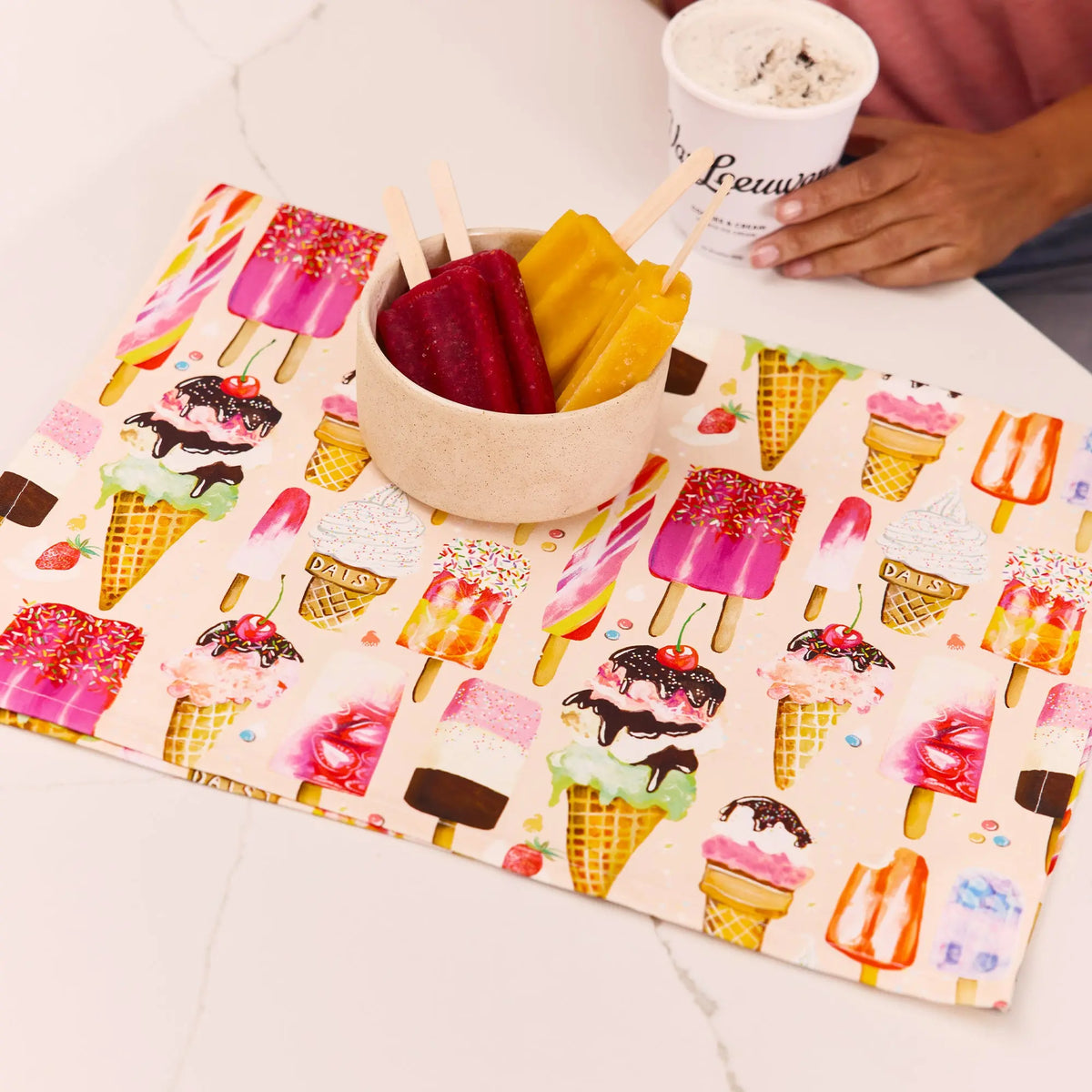 Cotton tea towel featuring a whimsical pattern of colorful ice cream cones and popsicles on a light pink background.