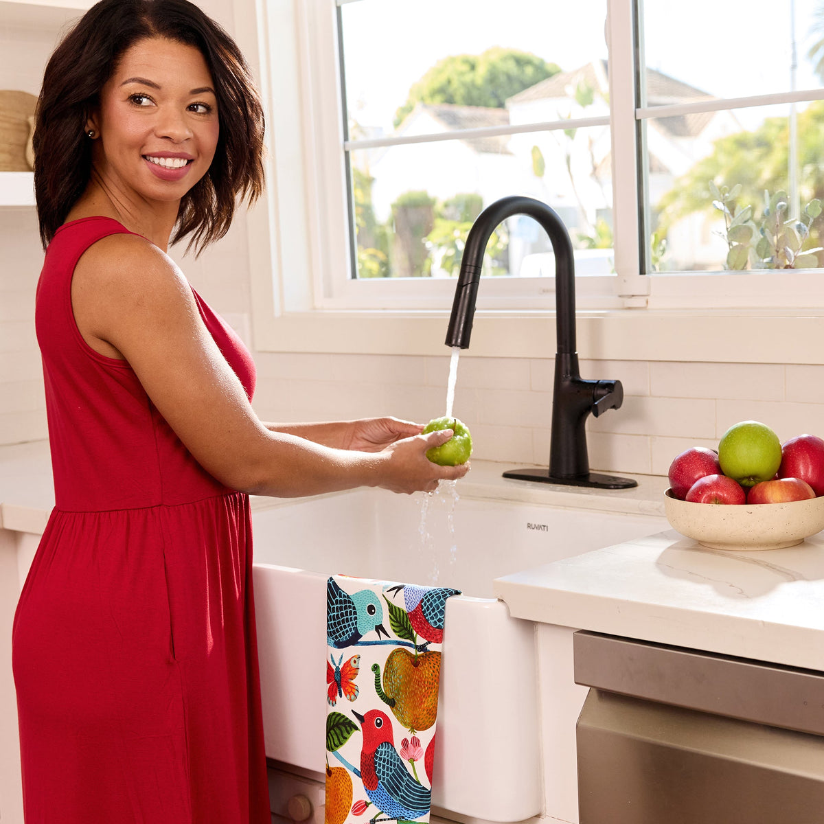 Woman in red dress washing a green apple; a cotton tea towel with colorful birds and apples hangs on the sink edge.
