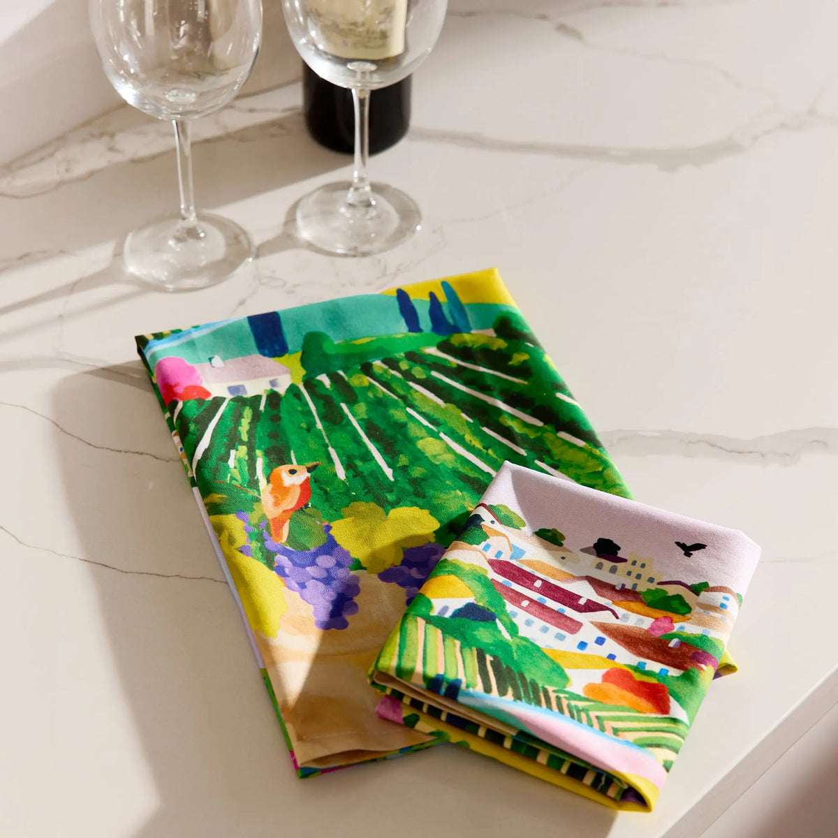 A cotton tea towel featuring a colorful, illustrated vineyard landscape with vines, grapes, and buildings on a counter.