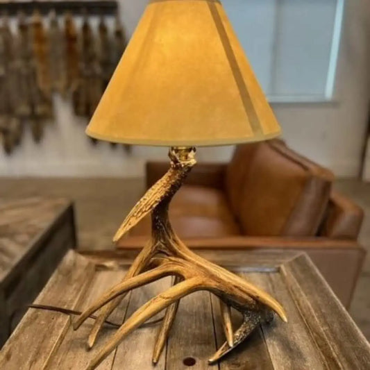 Table lamp with a rustic whitetail deer antler base and a tan, conical fabric shade on a wooden tabletop.