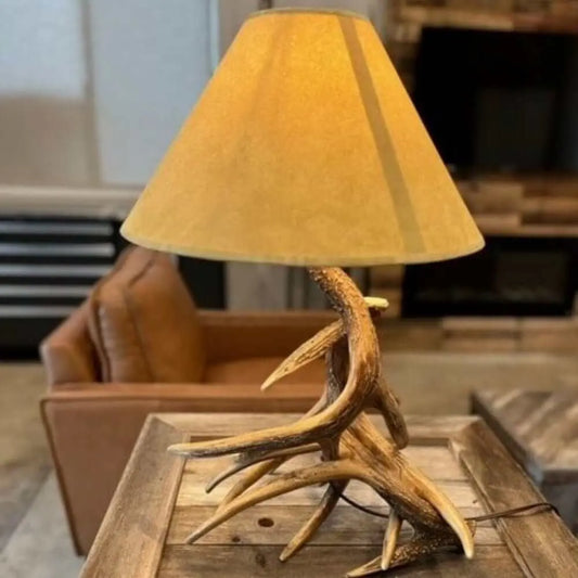 Rustic table lamp featuring a realistic whitetail antler base and a warm-toned, tapered fabric shade.