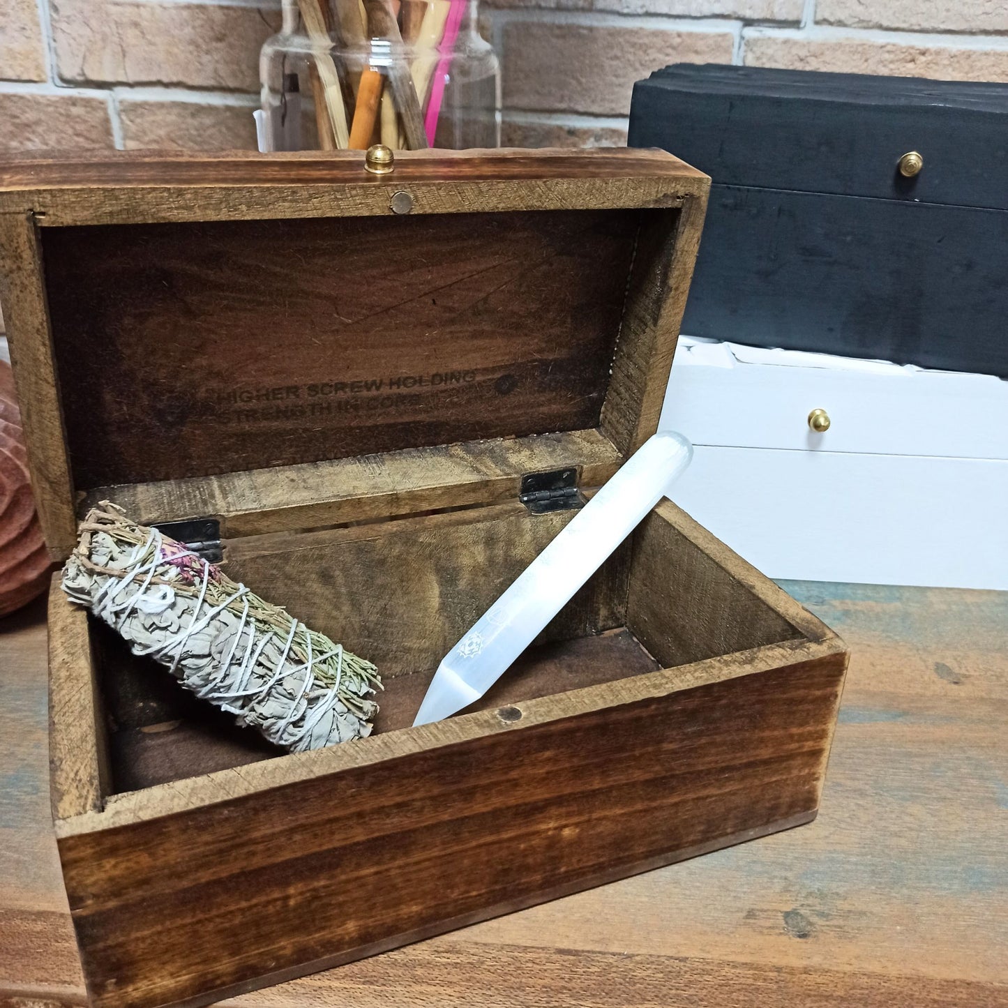 Caja de madera marrón con varita de cristal y palo de salvia dentro, ideal para almacenar elementos espirituales.
