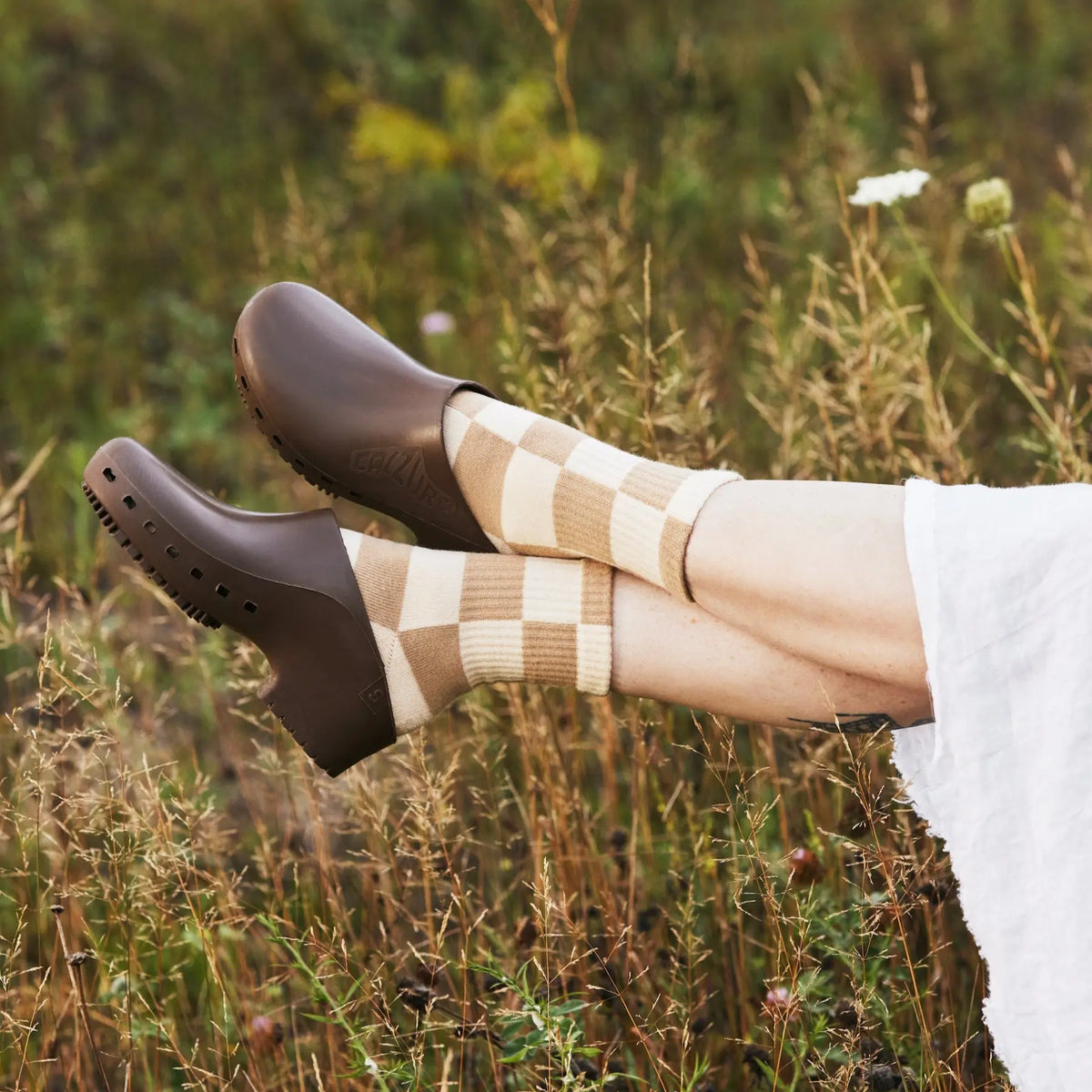Brown clogs with perforations paired with beige and brown checkered socks in a natural outdoor setting.