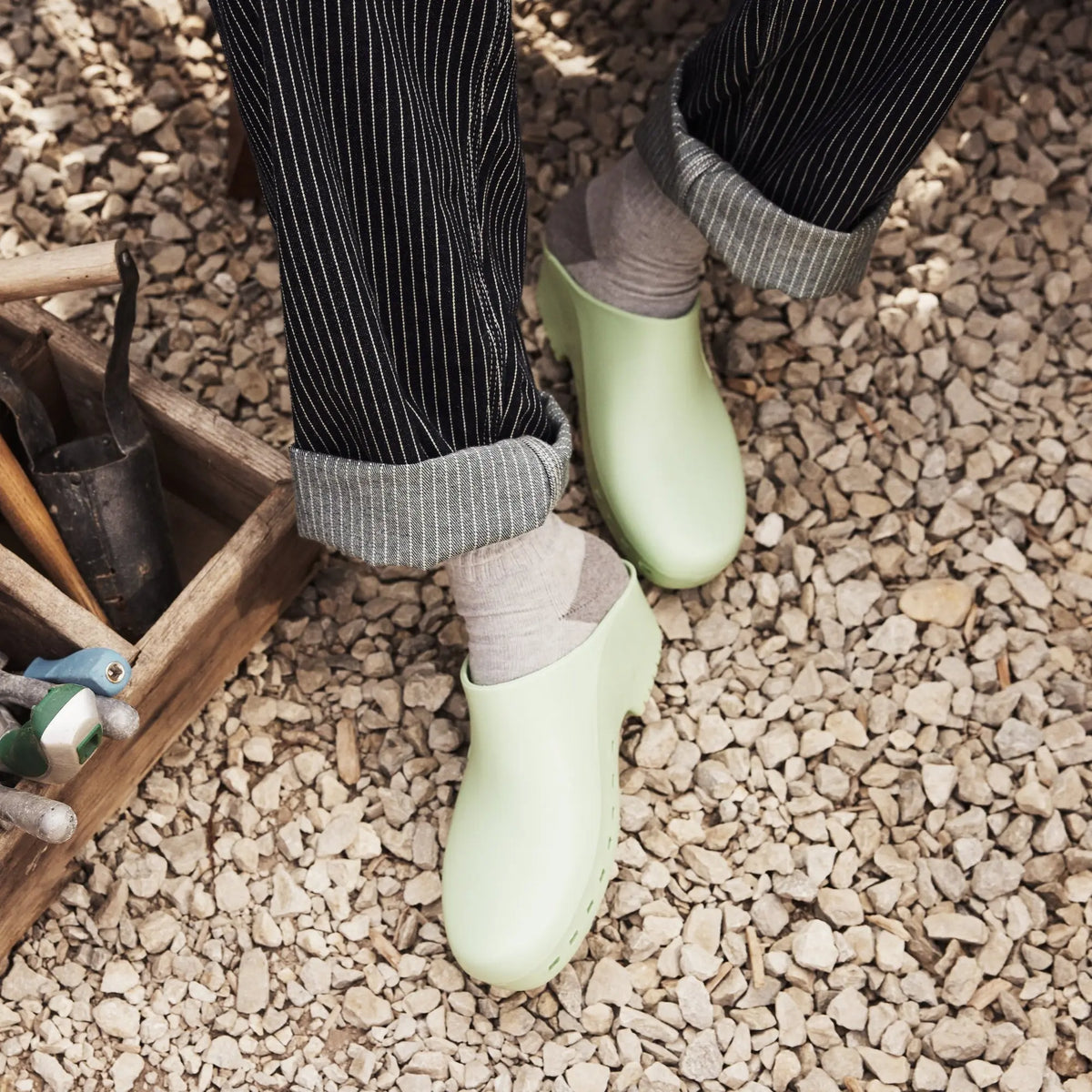 Light green rubber clogs worn with gray socks and striped pants, perfect for gardening or casual wear.