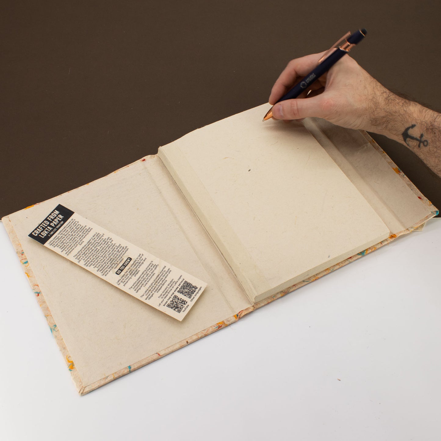 Handcrafted blank notebook with recycled paper, colorful patterned cover, and bronze pen on a neutral background.