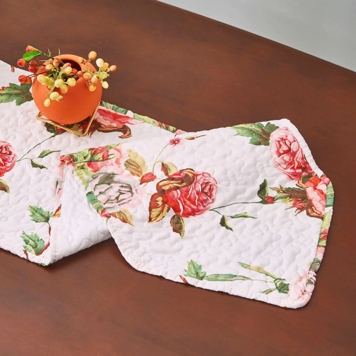 Quilted dining table runner with pink rose floral pattern on white fabric, featuring a scalloped edge on wooden table.