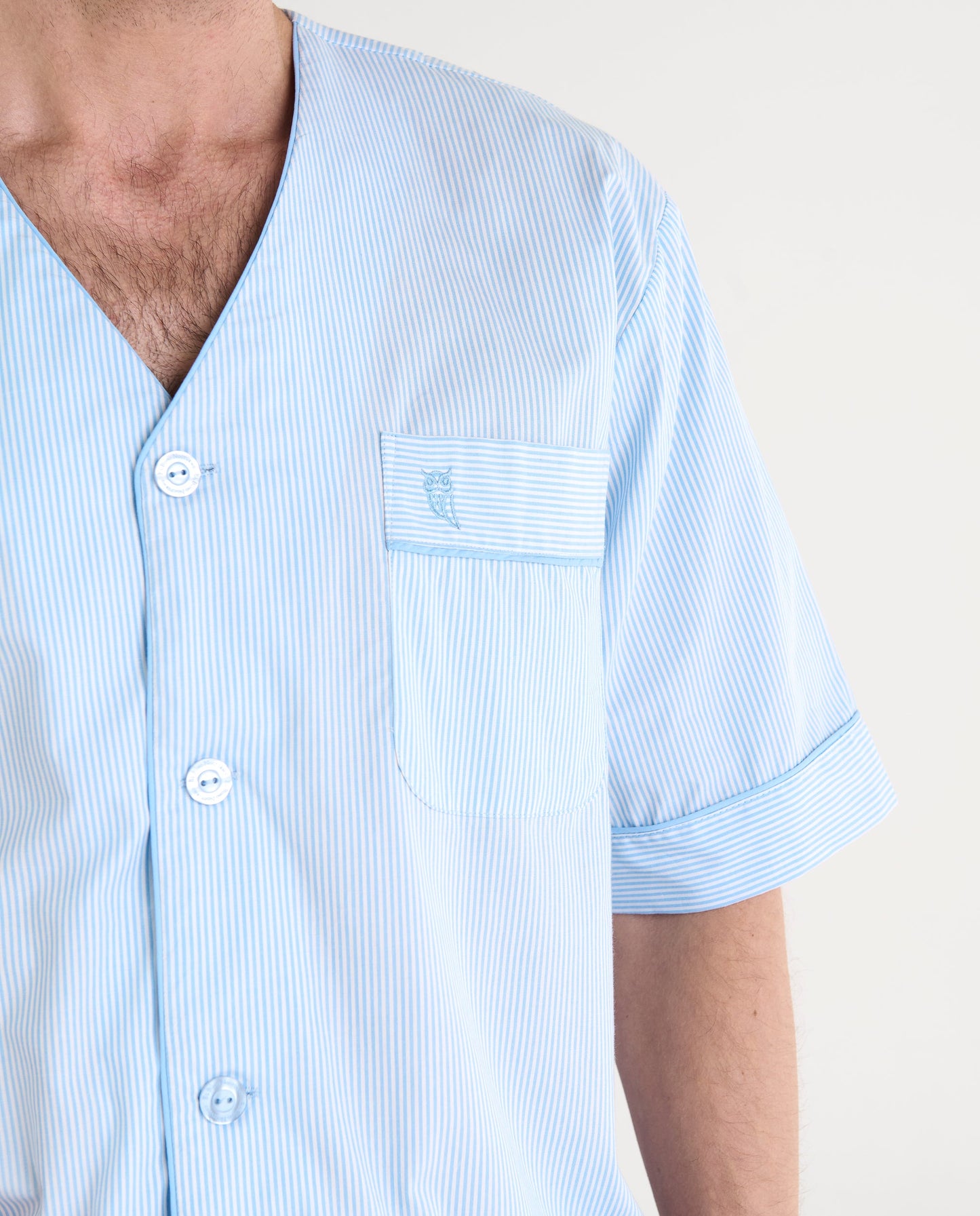Camisa de hombre azul claro a rayas, manga corta, con bolsillo en el pecho y botones frontales.