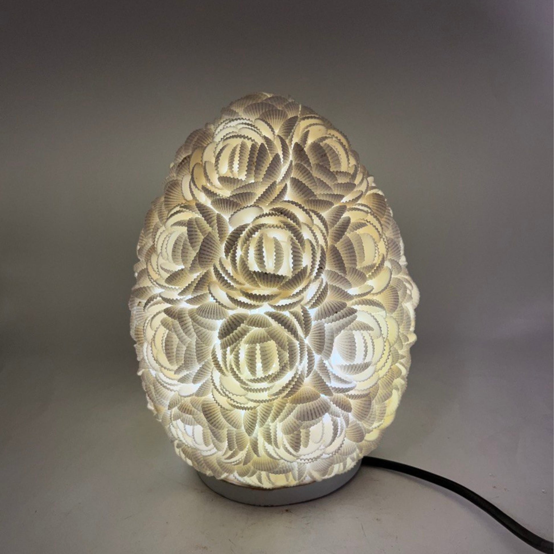 Lámpara boho ovalada de 20cm, hecha con conchas marinas blancas dispuestas en forma de rosas, con luz cálida interna.