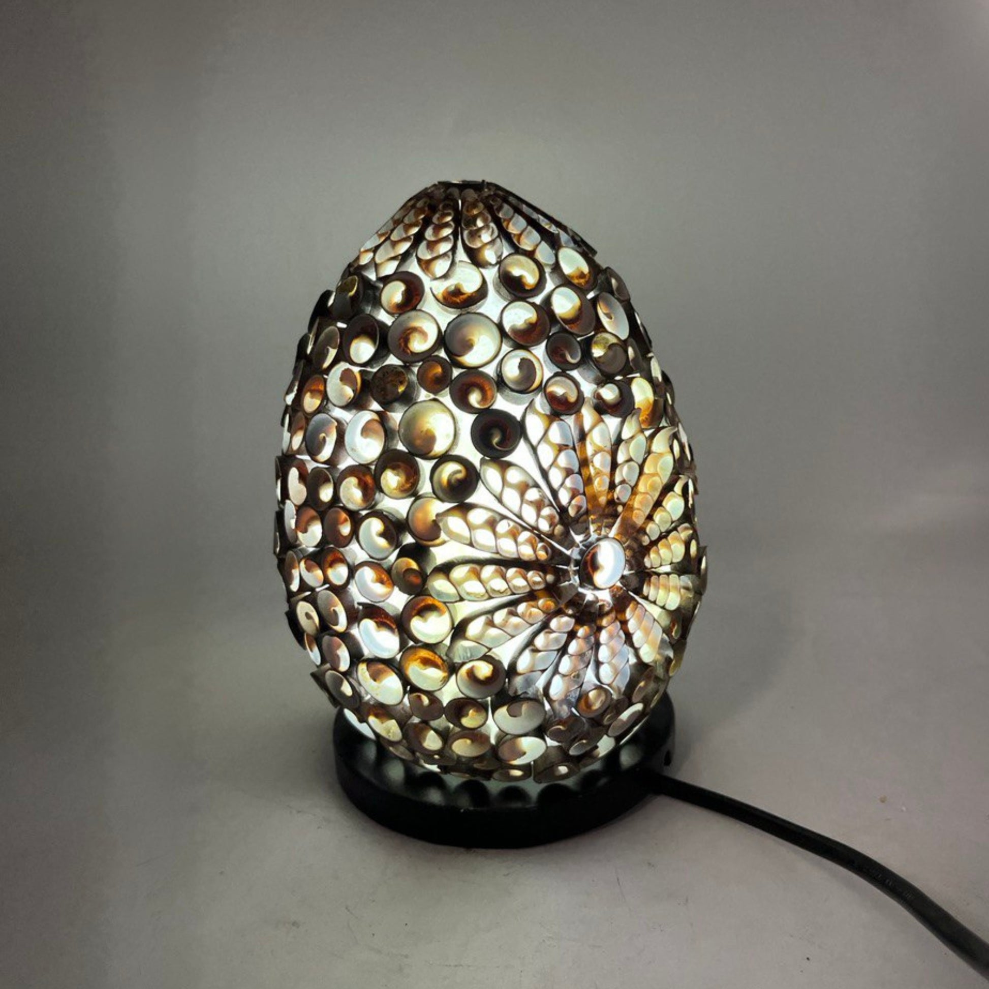 Lámpara boho ovalada de 20 cm, hecha de conchas marinas color chocolate con diseño de flor iluminada sobre base negra.
