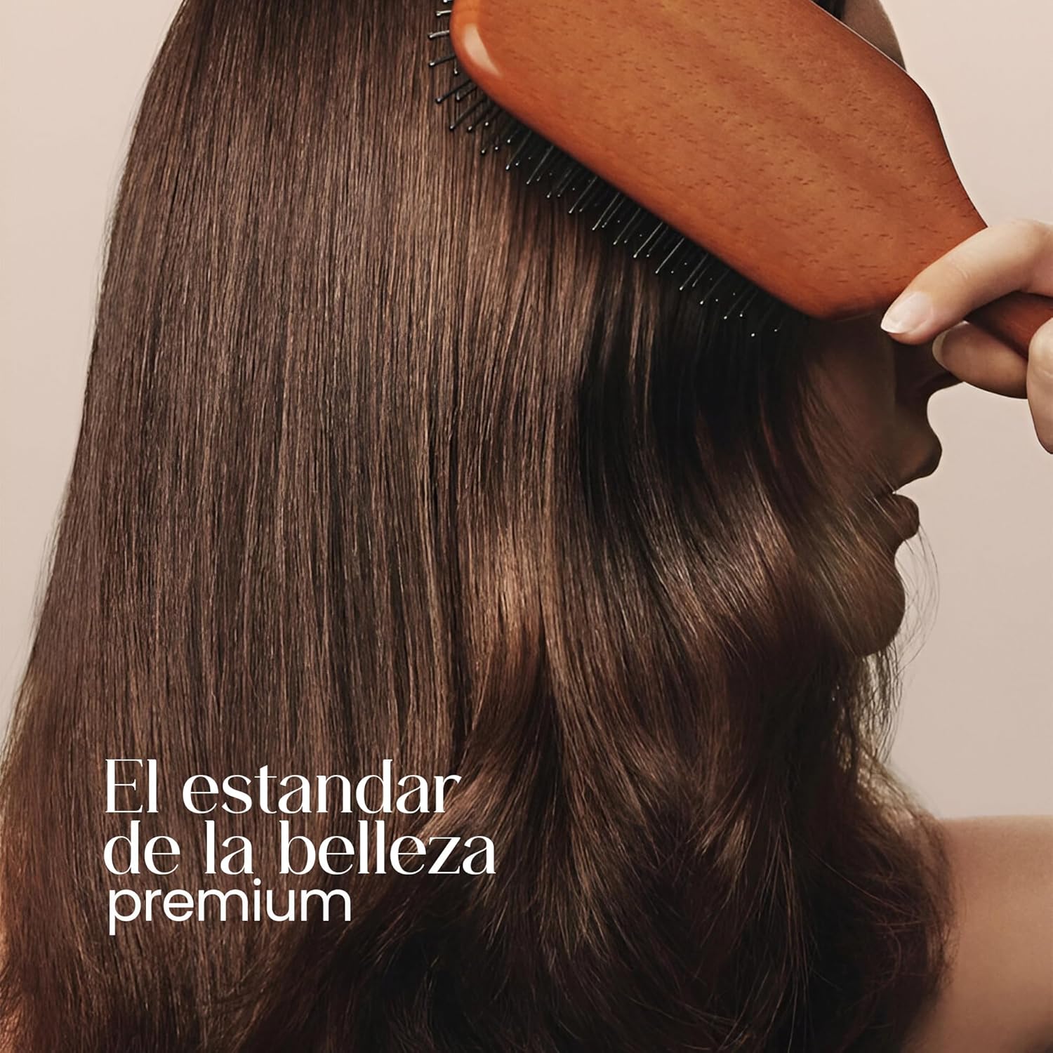 Mujer peinando su cabello castaño, liso y brillante con un cepillo de madera sobre fondo beige neutro.