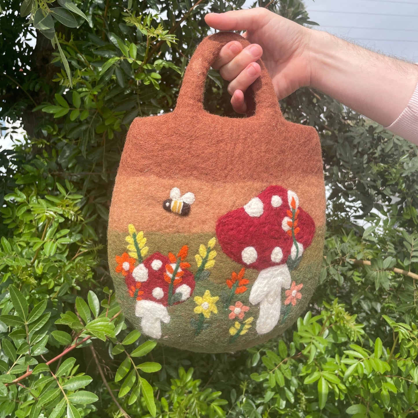 Bolsa de fieltro redondeada con diseño artesanal de setas, flores y abeja sobre fondo degradado de tonos tierra.