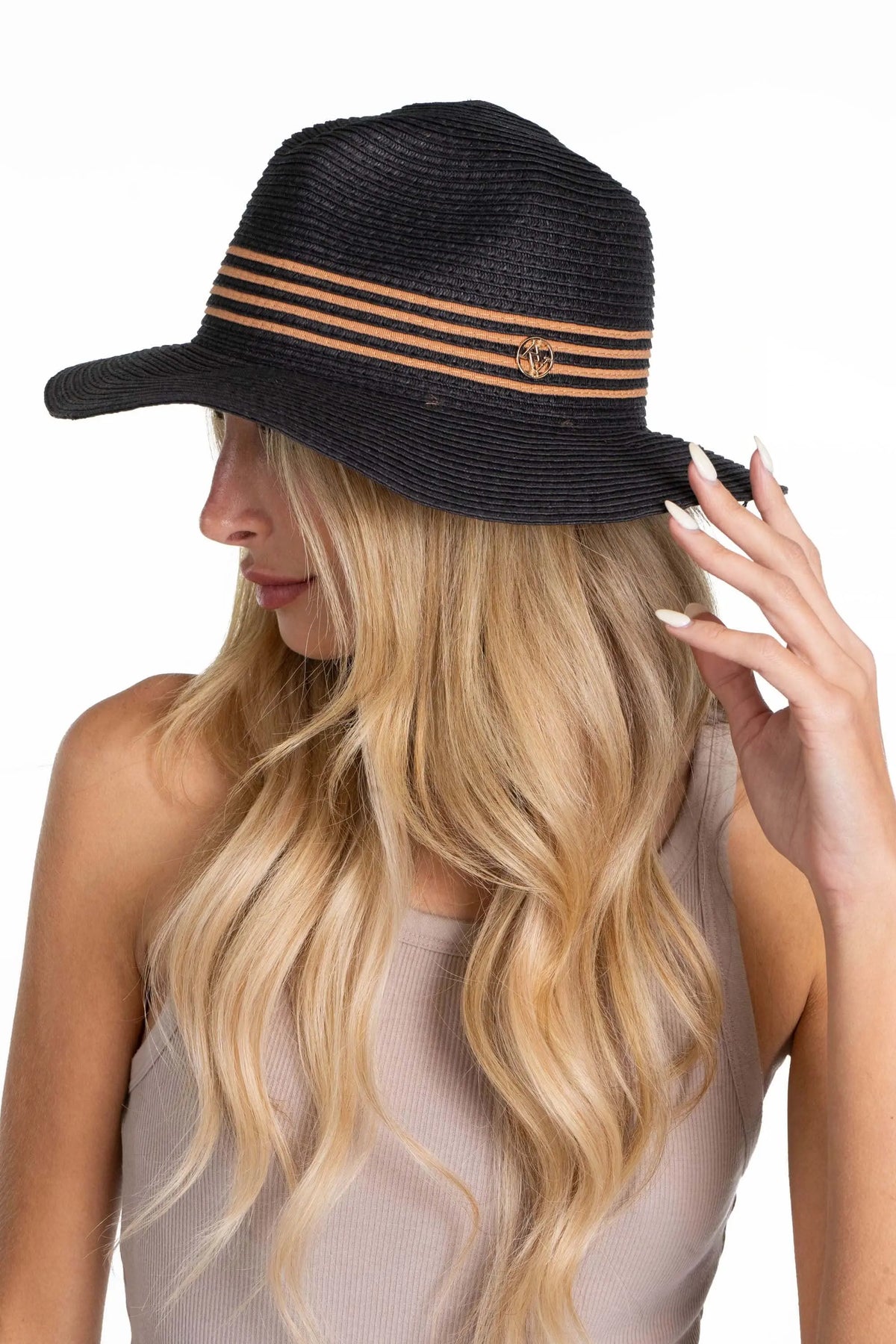 Black straw Panama hat with tan striped band and gold logo emblem, worn by a woman with long blonde hair.