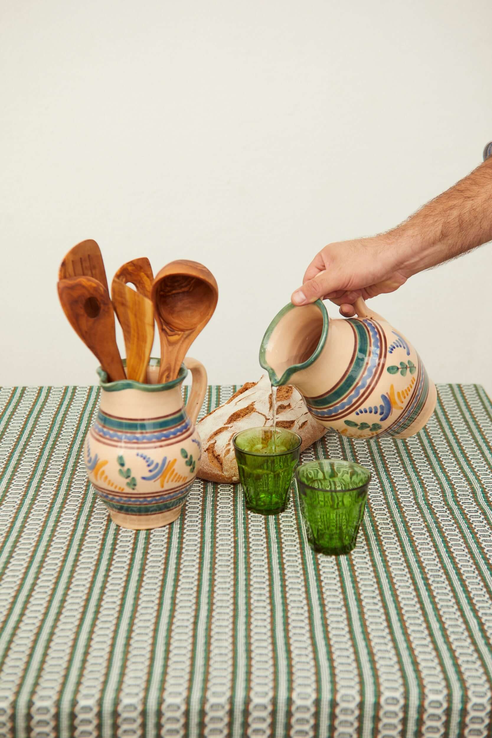 Jarra de cerámica artesanal beige con motivos pintados en azul y verde, vertiendo agua en vasos de cristal verde.