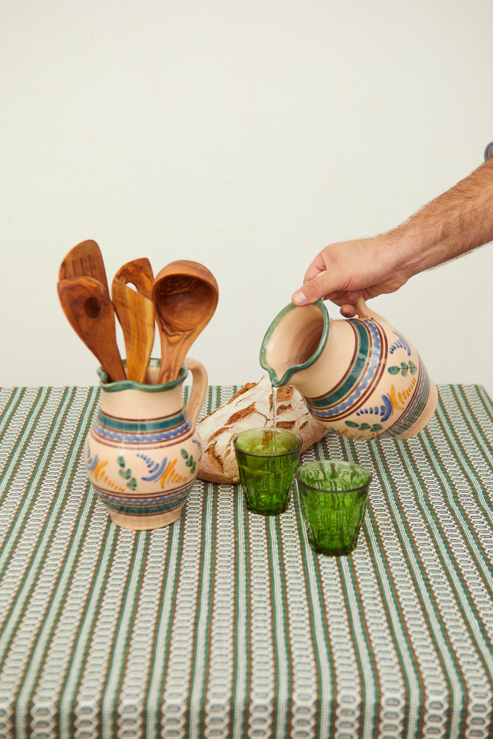 Jarras de cerámica pintadas a mano, cucharas de madera y vasos verdes sobre un mantel blanco y verde.