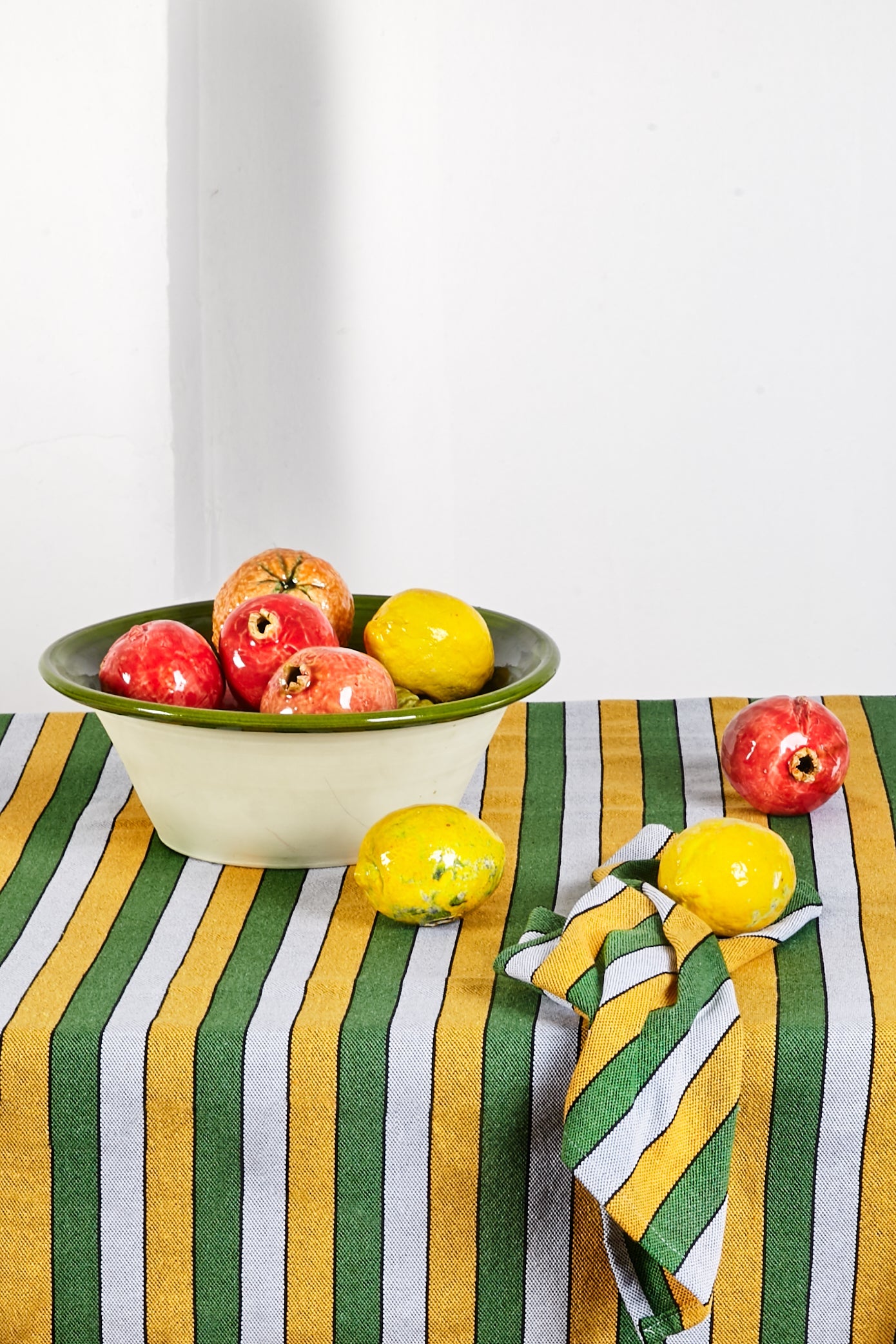 Tazón de cerámica verde y beige con manzanas rojas y limones amarillos sobre un mantel de rayas verdes y amarillas.