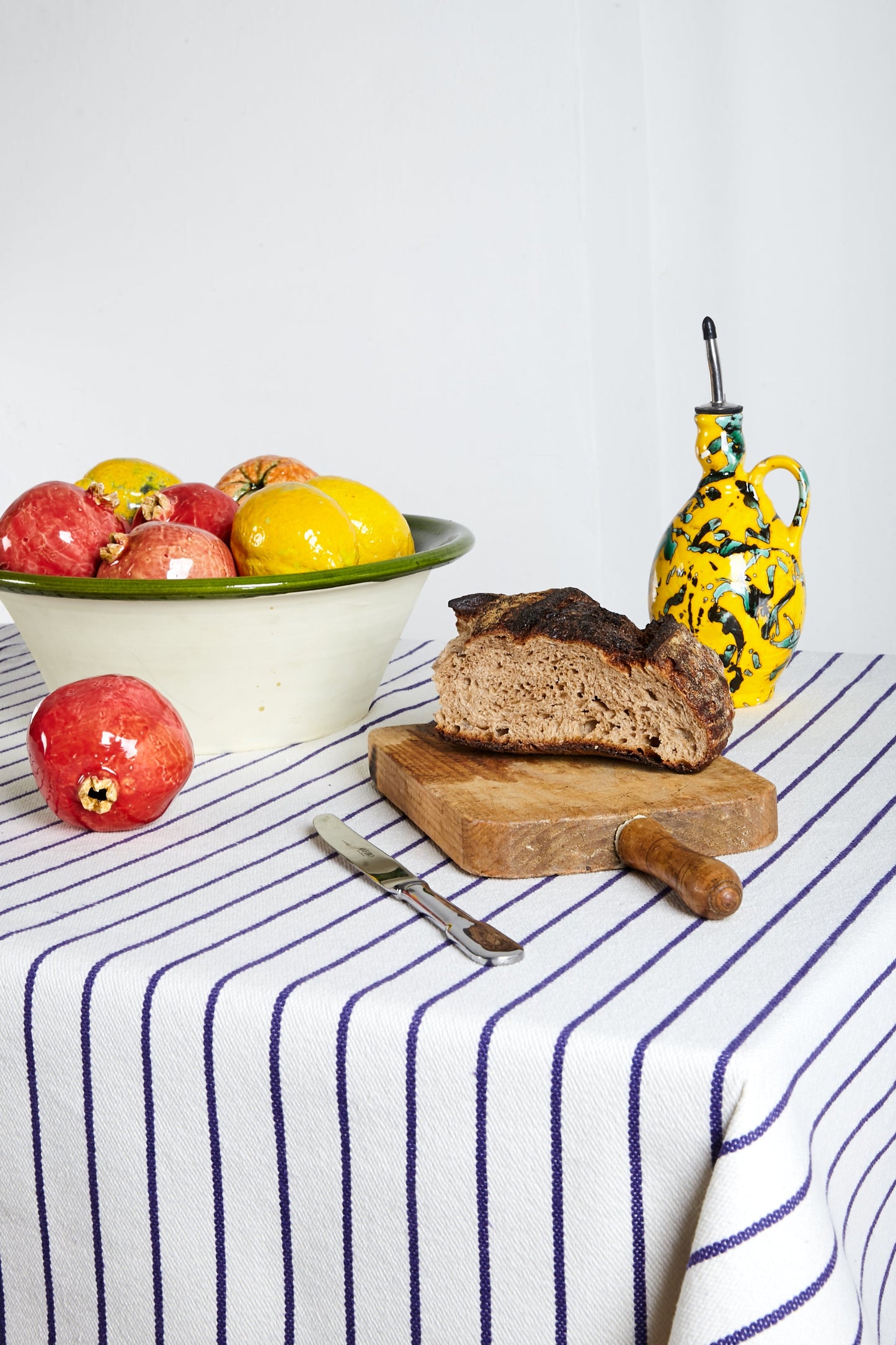 Pan marrón sobre una tabla de madera, con bowl de frutas coloridas y jarra amarilla decorativa, sobre manteles a rayas.