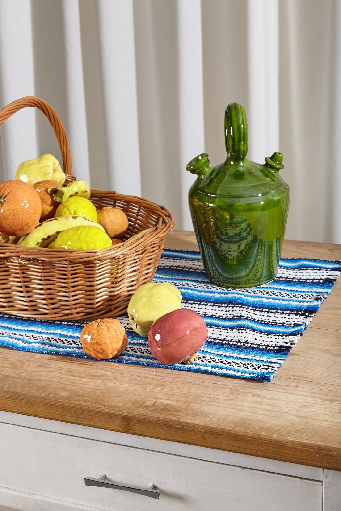 Cesta de mimbre con frutas y jarra verde sobre un camino de mesa azul y blanco en mesa de madera.