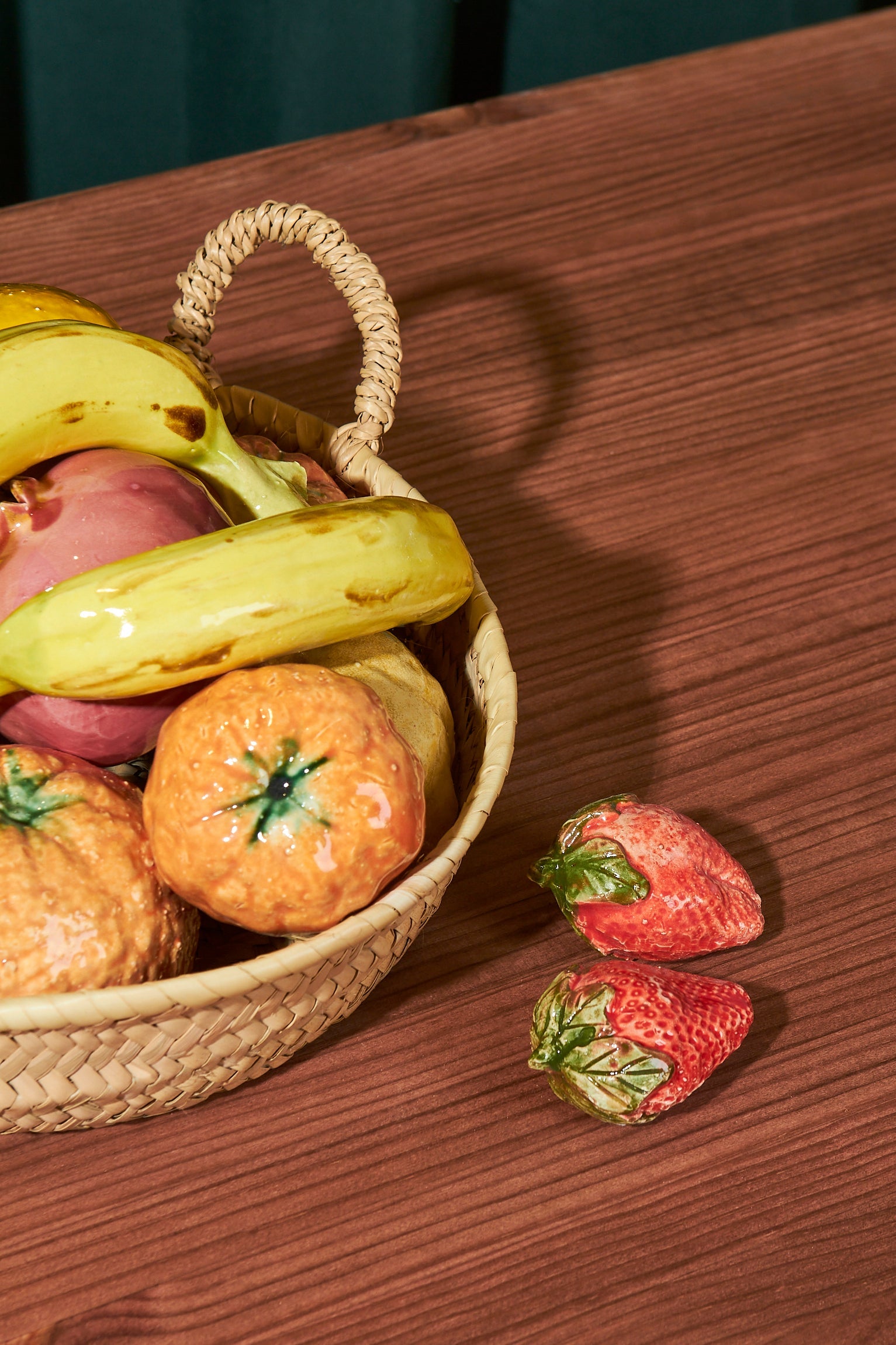 Cesta de mimbre con frutas de cerámica coloridas: plátanos, fresas y naranjas sobre mesa de madera.