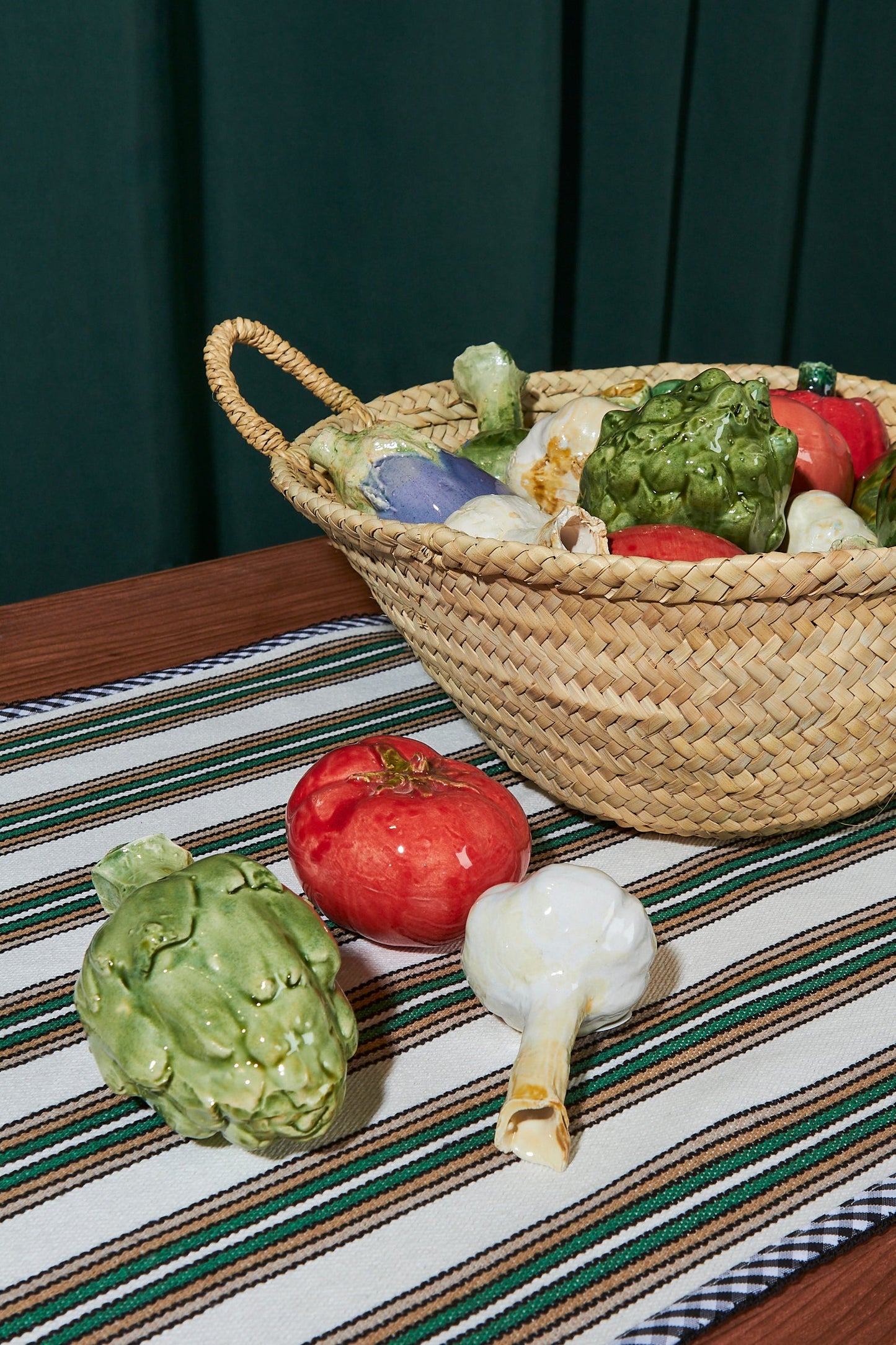 Cesta de mimbre con verduras de cerámica coloridas: tomate rojo, alcachofa verde y ajo blanco sobre un camino de mesa a rayas.