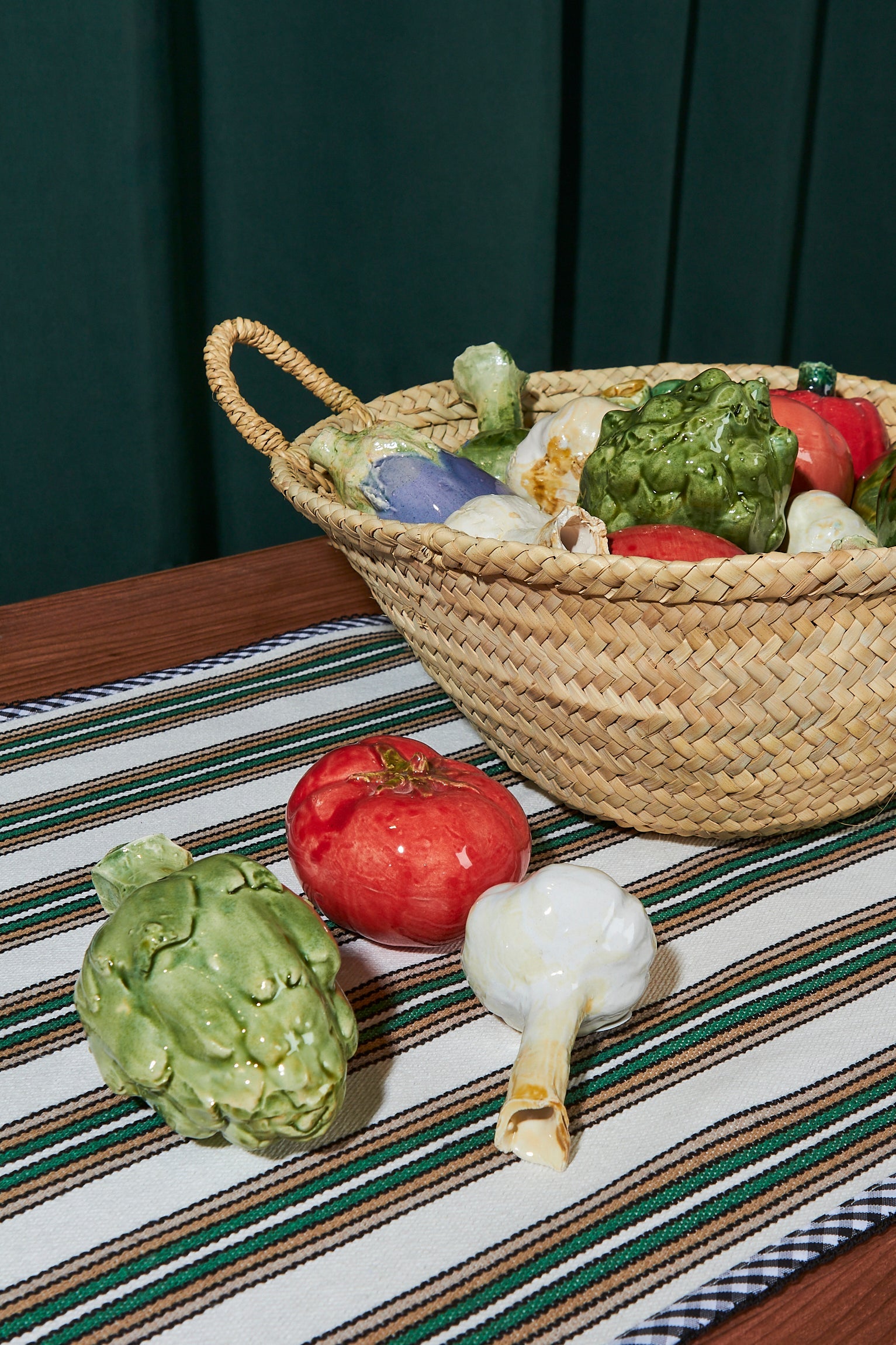 Cesta de mimbre con verduras de cerámica coloridas: tomate rojo, alcachofa verde y ajo blanco sobre un camino de mesa a rayas.
