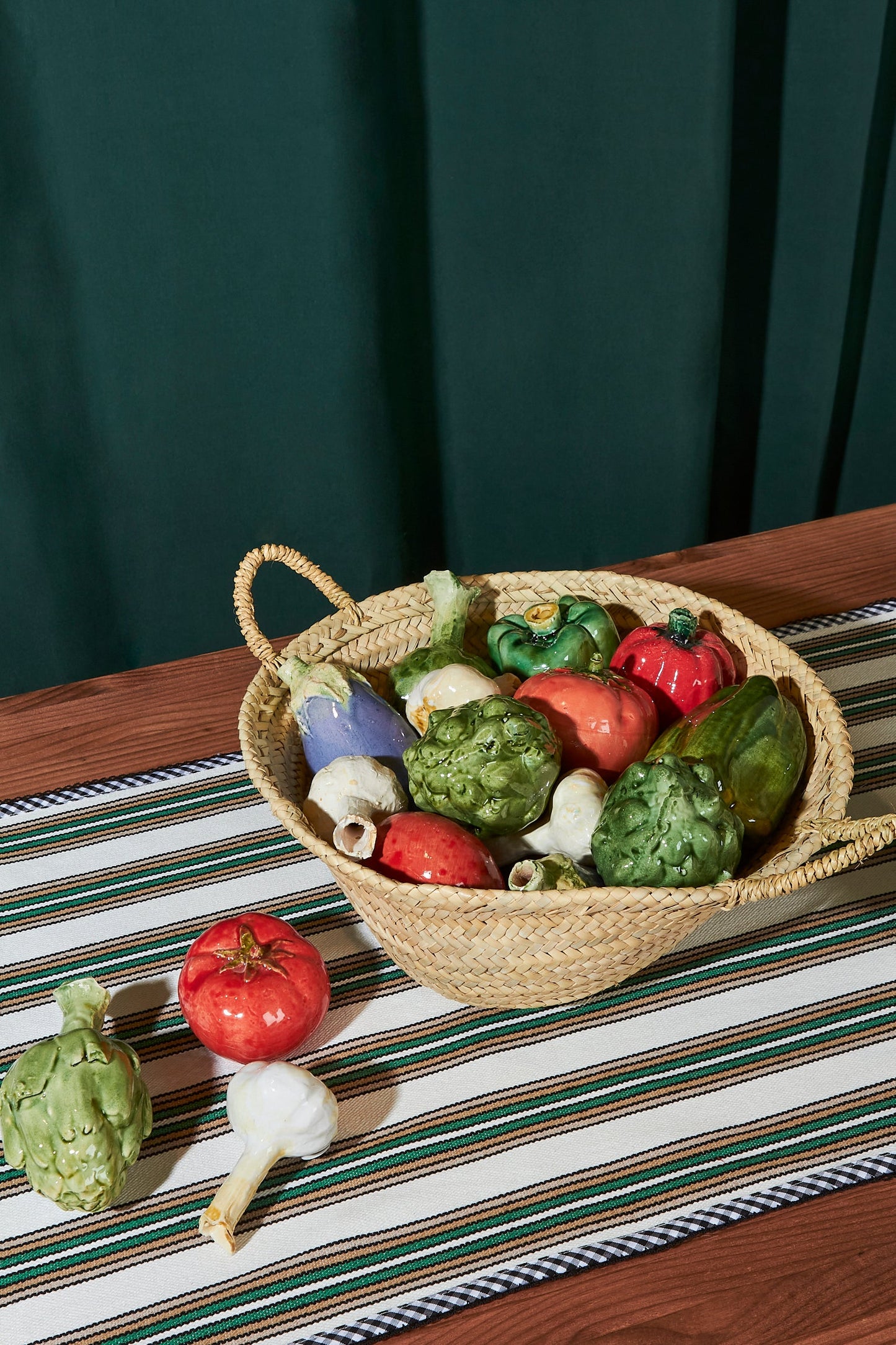 Cesta de mimbre con verduras de cerámica en colores rojo, verde y morado sobre un mantel a rayas.