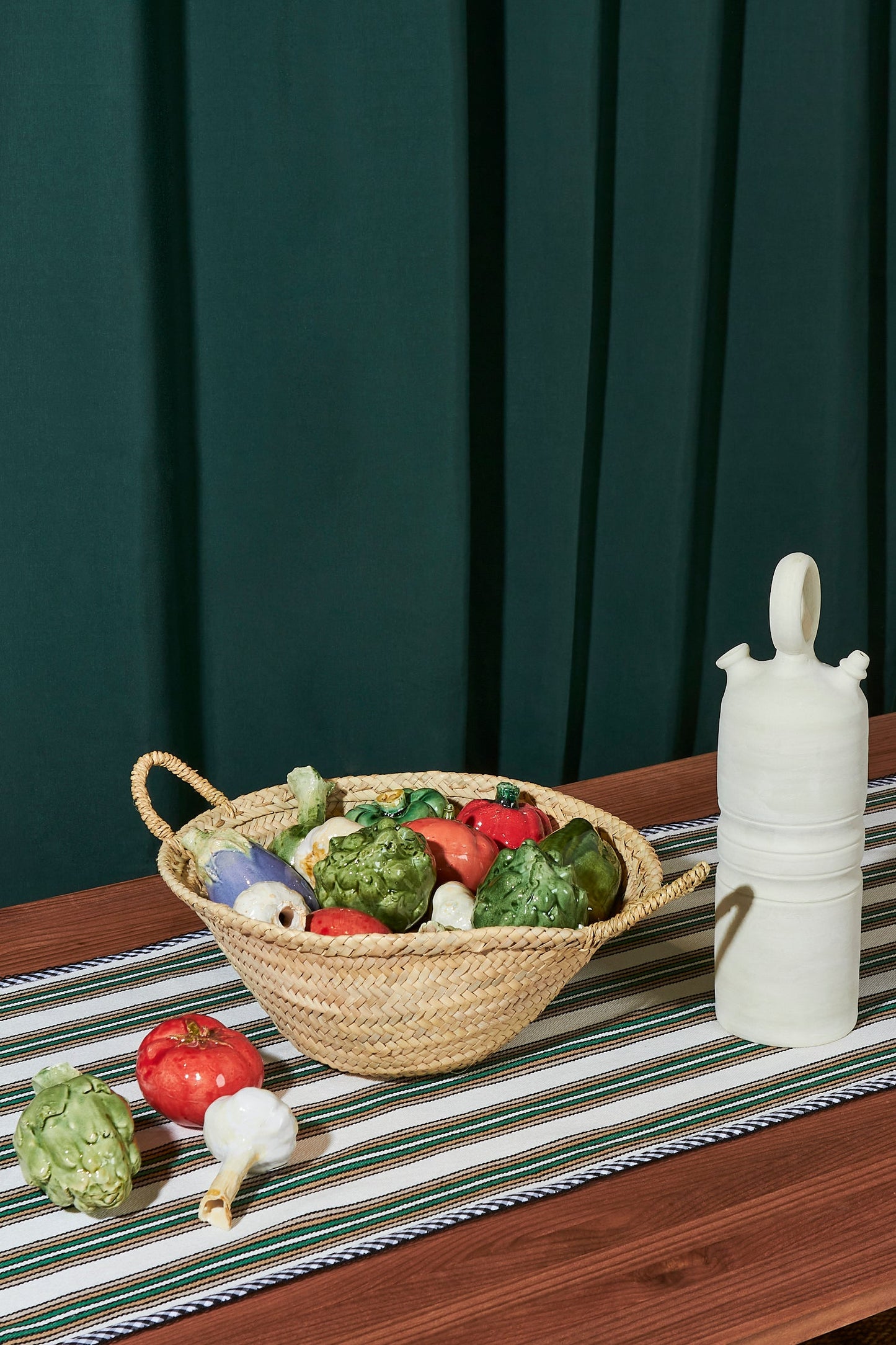 Cesta de mimbre con verduras decorativas de colores vivos sobre mantel a rayas, fondo verde oscuro.