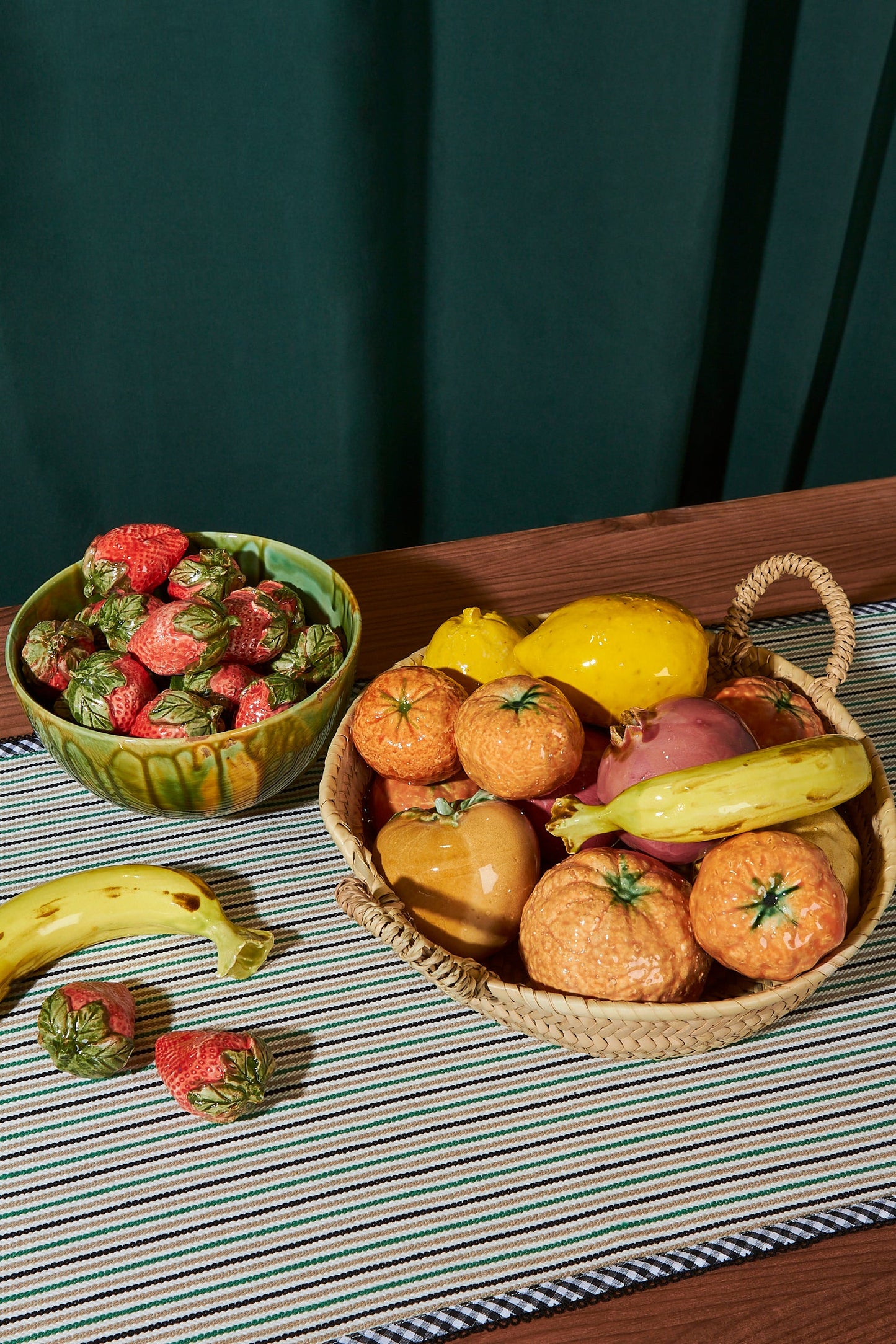 Frutas de cerámica en tonos naranja y amarillo, con fresas rojas en un tazón verde, sobre un mantel de rayas.