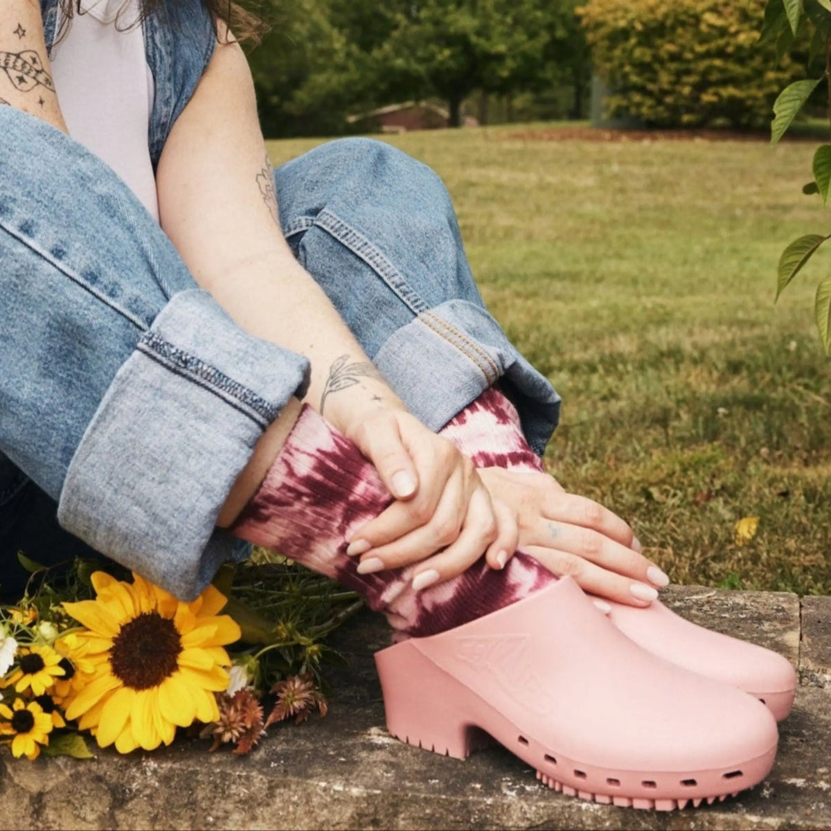 Bubblegum pink rubber clogs, hole-free design, worn with tie-dye socks, sitting on stone with sunflowers.