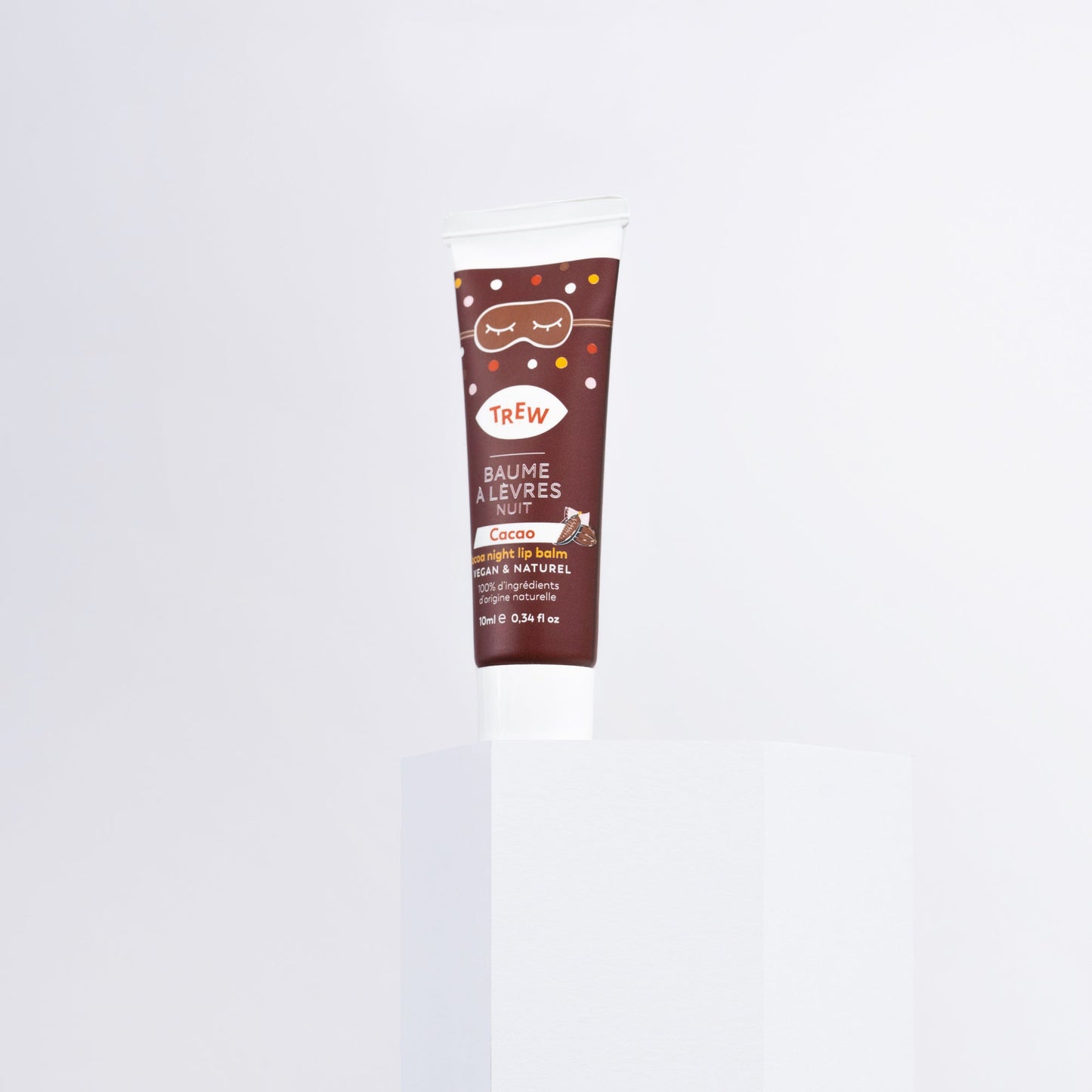 A brown cocoa lip mask tube with a sleep mask illustration, standing on a white pedestal against a light background.