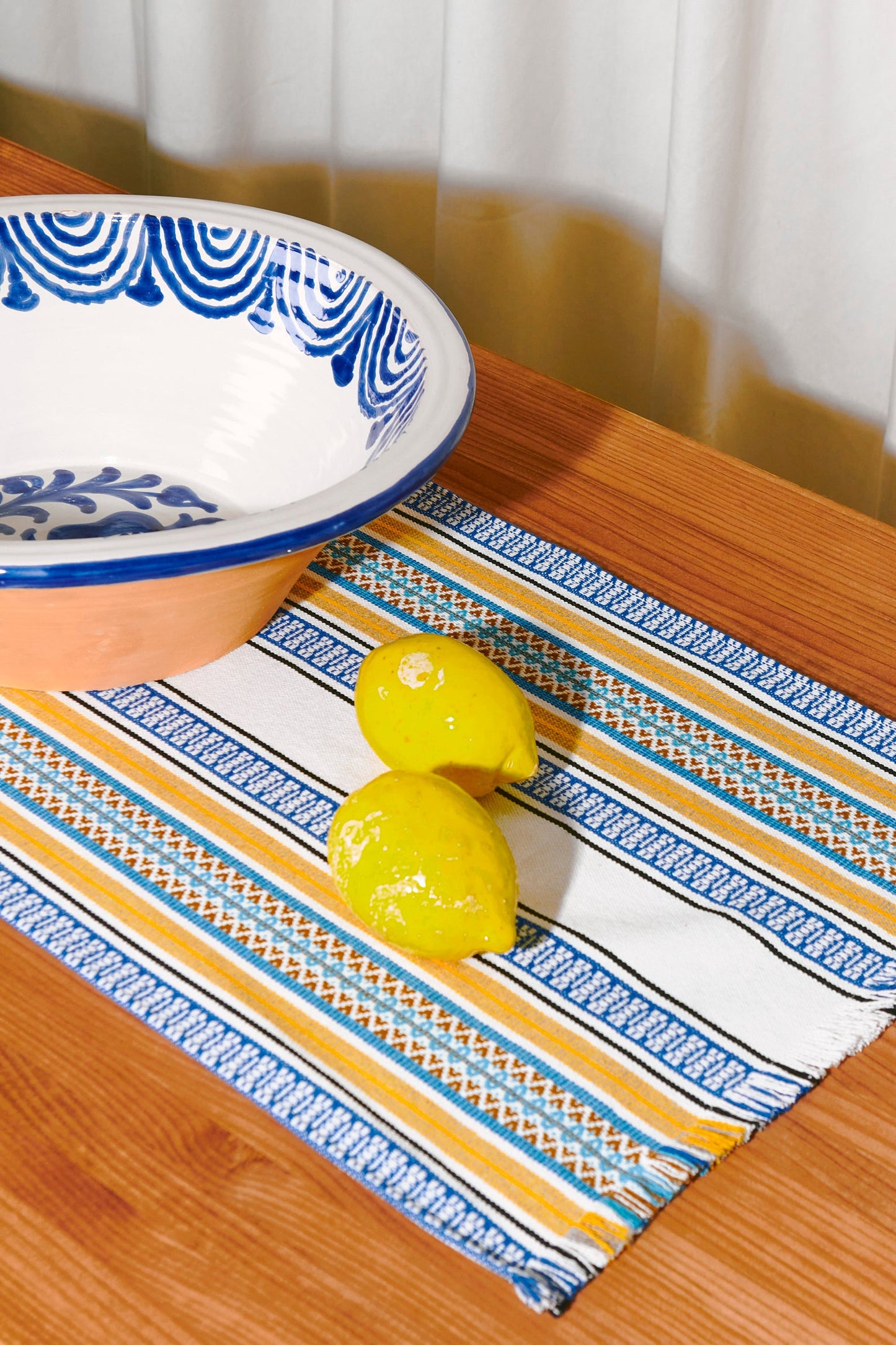 Mantel de rayas azules y amarillas con limones sobre una mesa de madera, junto a un tazón de cerámica artesanal.